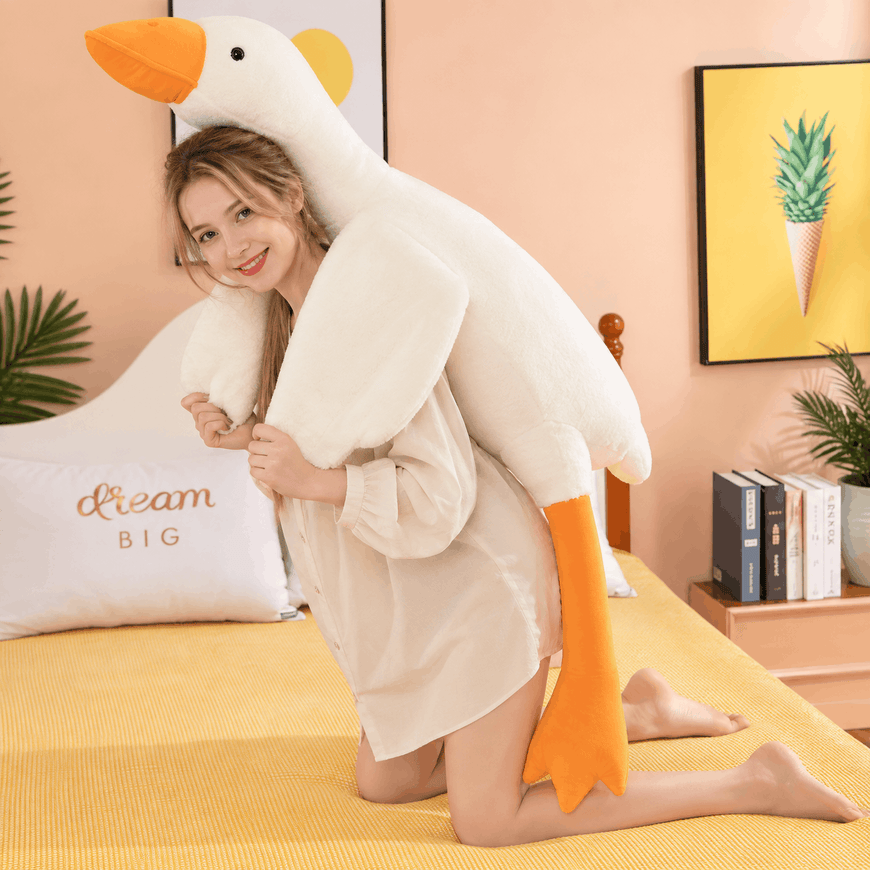 A woman kneeling on a yellow bed in a bedroom, holding a large plush goose toy. The goose is off-white with a bright orange beak, feet, and inner ear details.