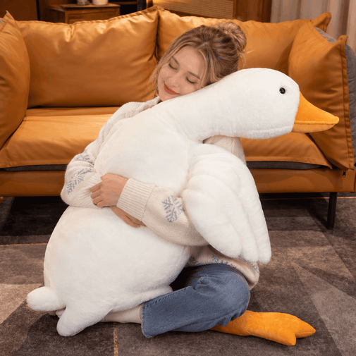 A woman hugging a very large white plush goose toy in a cozy living room setting with an orange-brown sofa in the background.