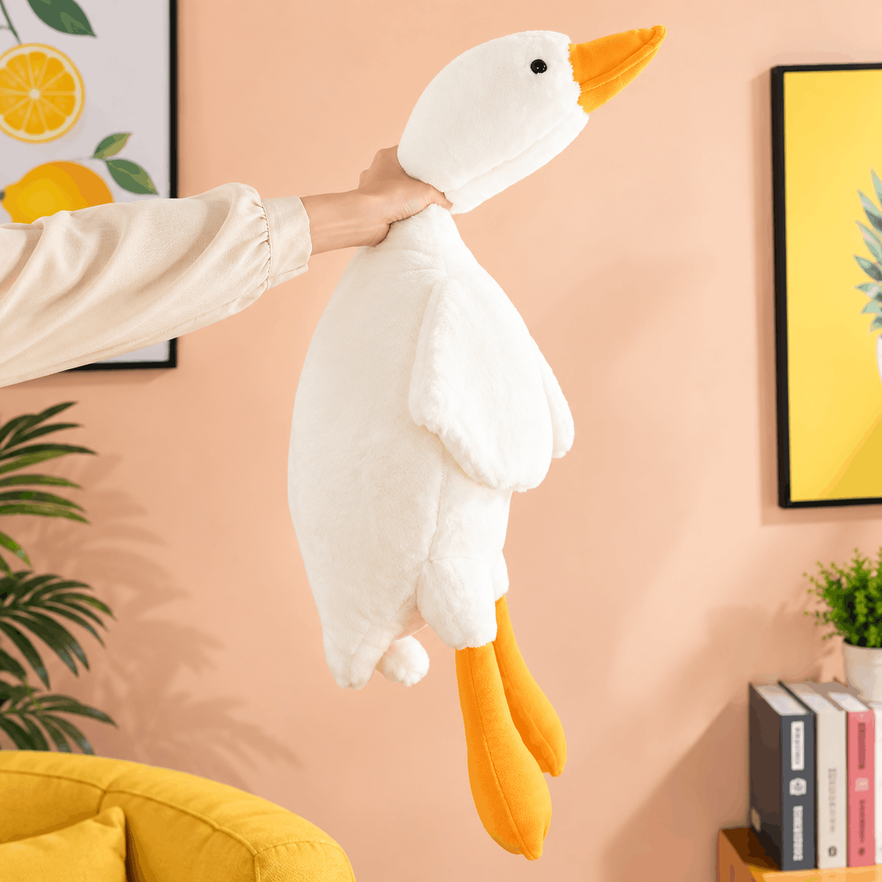 A person holds up a large white plush goose toy with orange beak and legs in a brightly lit room with a peach wall, framed art, plants, and a yellow chair.