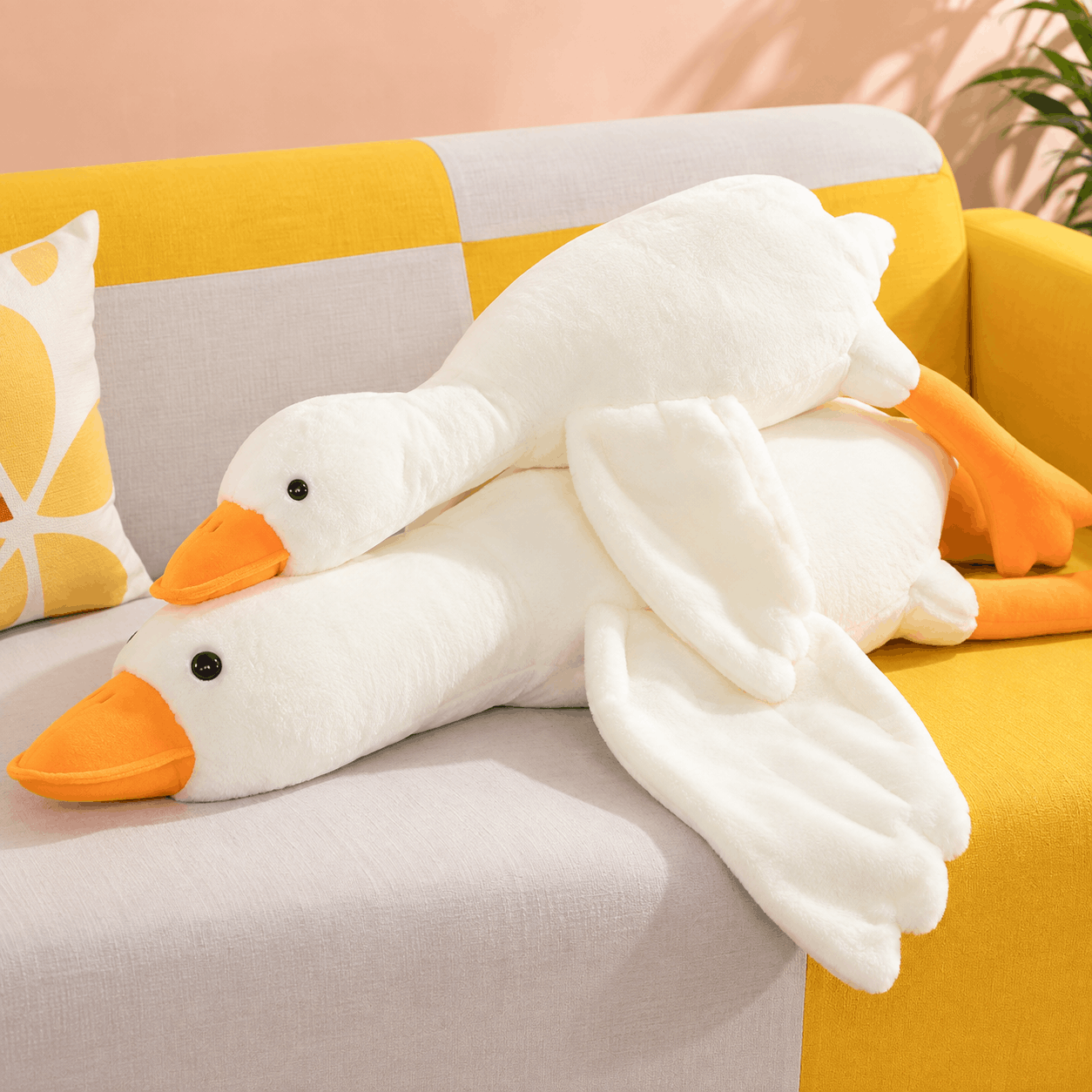 Two white plush goose toys with orange beaks and feet resting on a yellow and gray sofa with a patterned pillow.