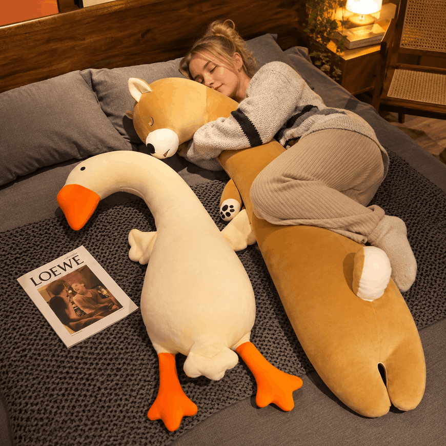 A cozy bedroom scene with a woman sleeping on a bed surrounded by plush toys, including a large tan animal-shaped pillow, a white goose plush with orange feet, and a fox plush. A Loewe magazine lies on the bed beside them.