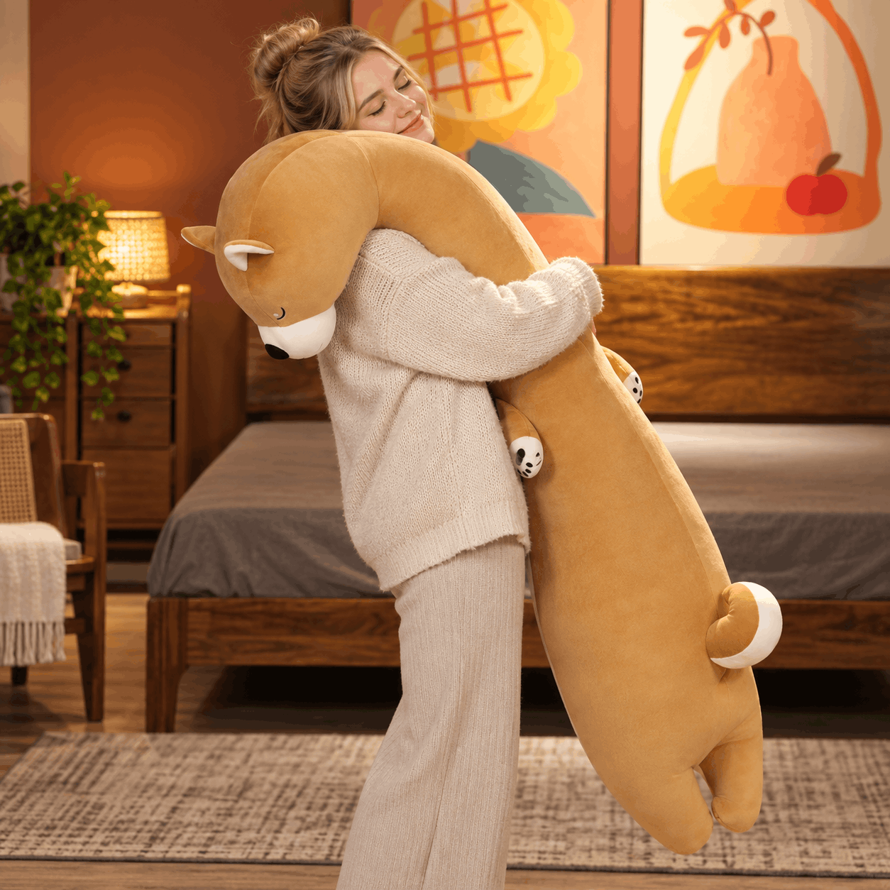 A woman in a cozy bedroom hugging a very large plush animal toy shaped like a long-necked fox or beaver in warm tan and brown tones with small white accents.