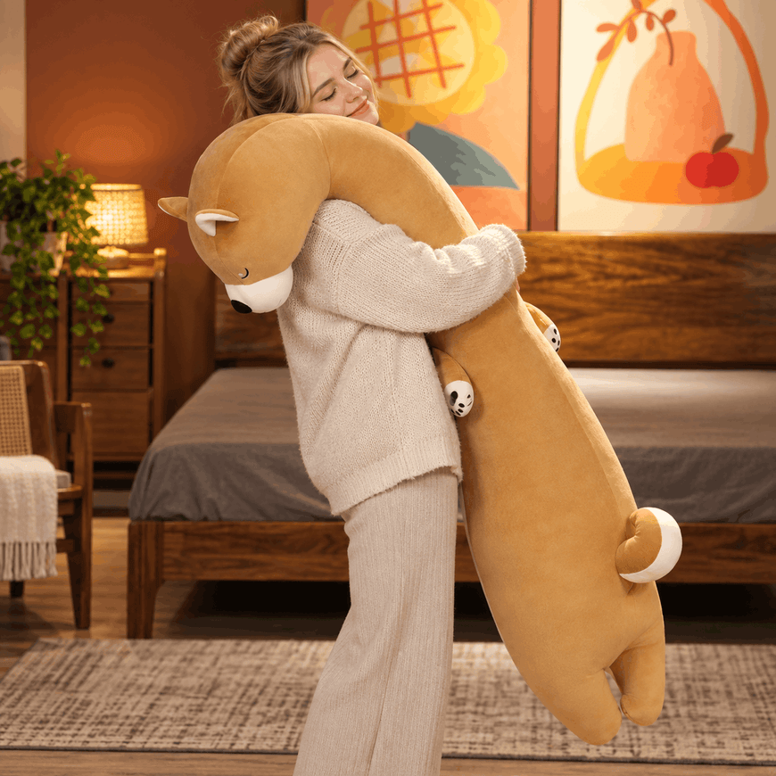 A woman in a cozy bedroom hugging a very large plush animal toy shaped like a long-necked fox or beaver in warm tan and brown tones with small white accents.