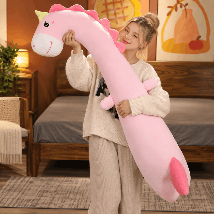 A woman in a cozy bedroom setting holds a large, long-necked pink dinosaur plush toy. The plushie has a white snout, pink horns, and a pink tail, and the woman is smiling while holding it.