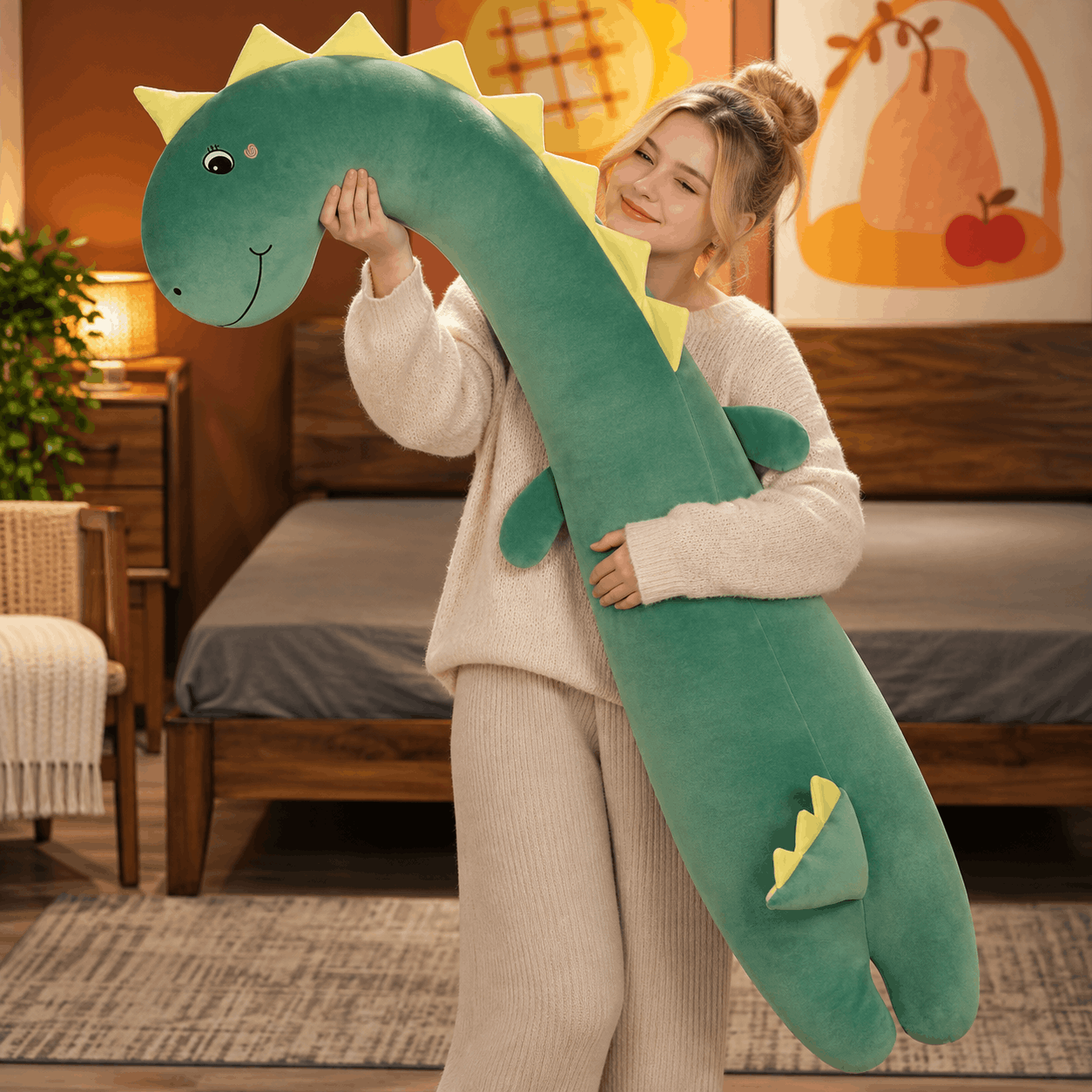 A woman in a cozy cream sweater and matching pants holds a long, plush green dinosaur toy with yellow spikes and a smiling face in a warmly lit bedroom.
