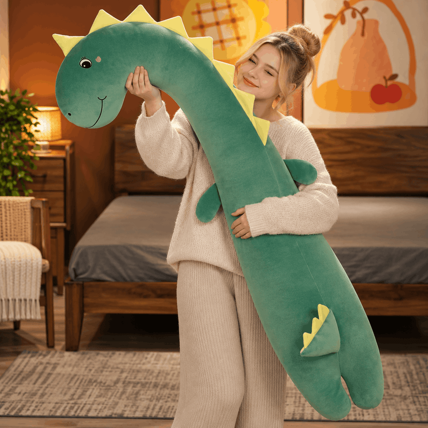 A woman in a cozy cream sweater and matching pants holds a long, plush green dinosaur toy with yellow spikes and a smiling face in a warmly lit bedroom.