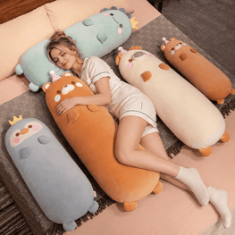 A woman lies on a bed surrounded by four large, cartoonish, animal-shaped plush pillows in pastel colors including blue, orange, cream, and gray. The pillows are decorated with cute faces, small ears, and tiny party hats.