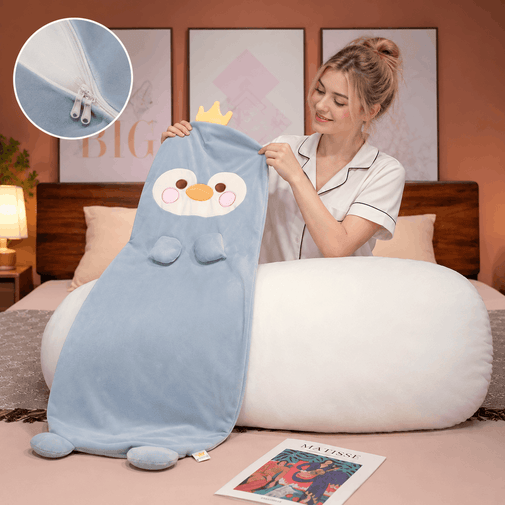 A woman in a bedroom holds a light blue plush penguin blanket with a white belly patch, pink cheeks, and a small yellow crown. The scene includes a large white oval pillow, framed wall art, a bed headboard, and a circular inset showing a close-up of the zipper detail.