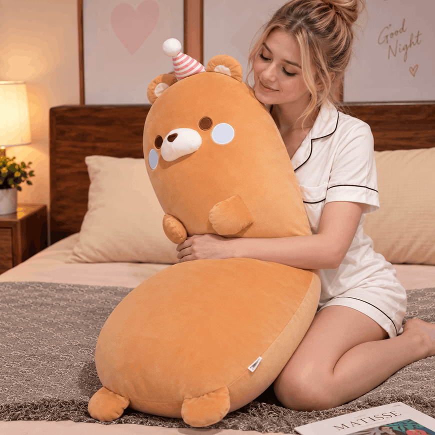 A woman in white pajamas hugs a large orange plush bear pillow on a bed in a cozy bedroom setting.