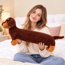 A woman sitting on a bed holds a long, plush dachshund-shaped pillow or toy. The product is brown with tan accents on the muzzle, paws, and tail.