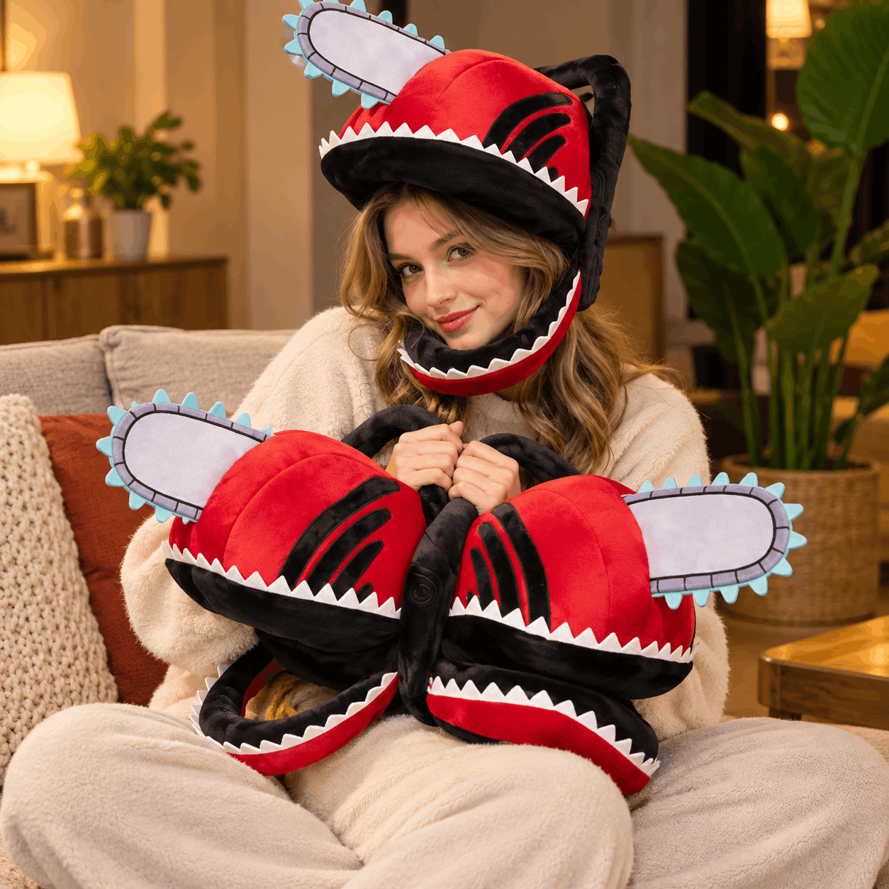 A woman wearing a red and black chainsaw-themed plush hood sits on a couch holding a matching plush pillow in a cozy living room setting.
