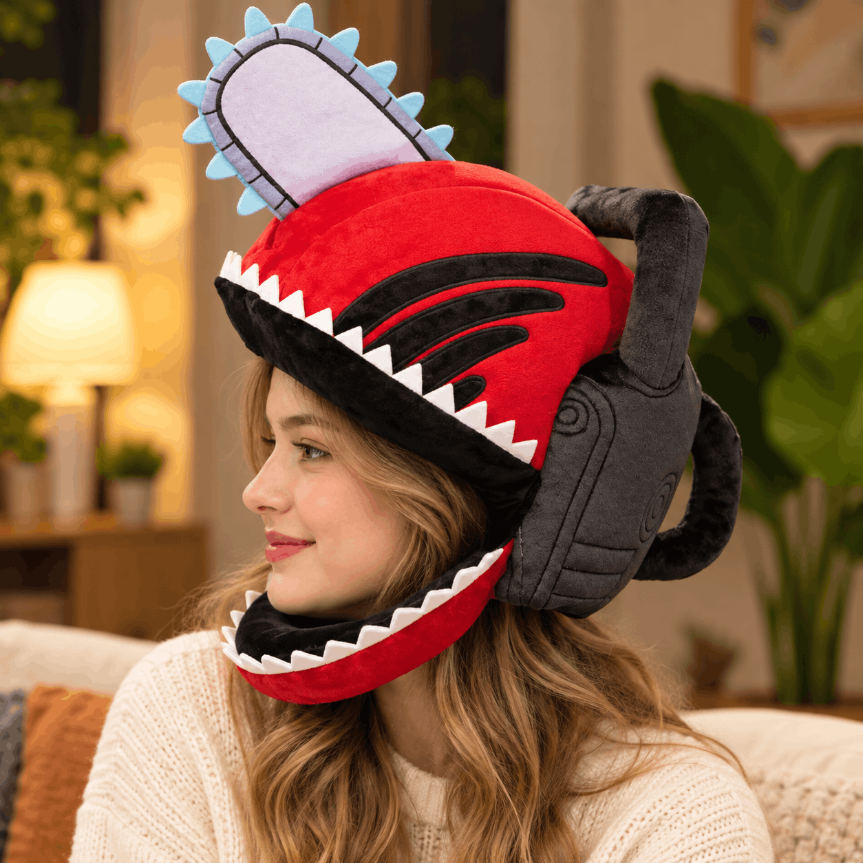 A woman wearing a plush chainsaw hat in a cozy living room setting. The hat is bright red with black details, white teeth, and a gray handle, with a light blue and white blade.