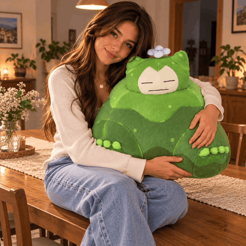 A woman sitting on a wooden dining table in a cozy, warmly lit room, holding a large green plush toy with a cartoon face and a small blue flower on its head.