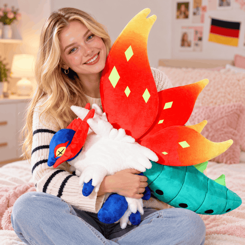 A smiling woman sits on a bed holding a large plush toy with a red and yellow flame-shaped back, a white body, blue accents, and a teal tail. The room has a cozy bedroom setting with a bed, lamp, and framed pictures in the background.