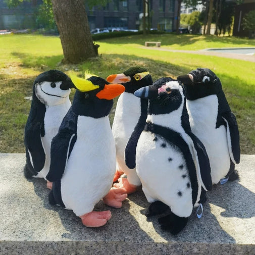 Five plush penguins sitting on a stone ledge with a grassy area and trees in the background.