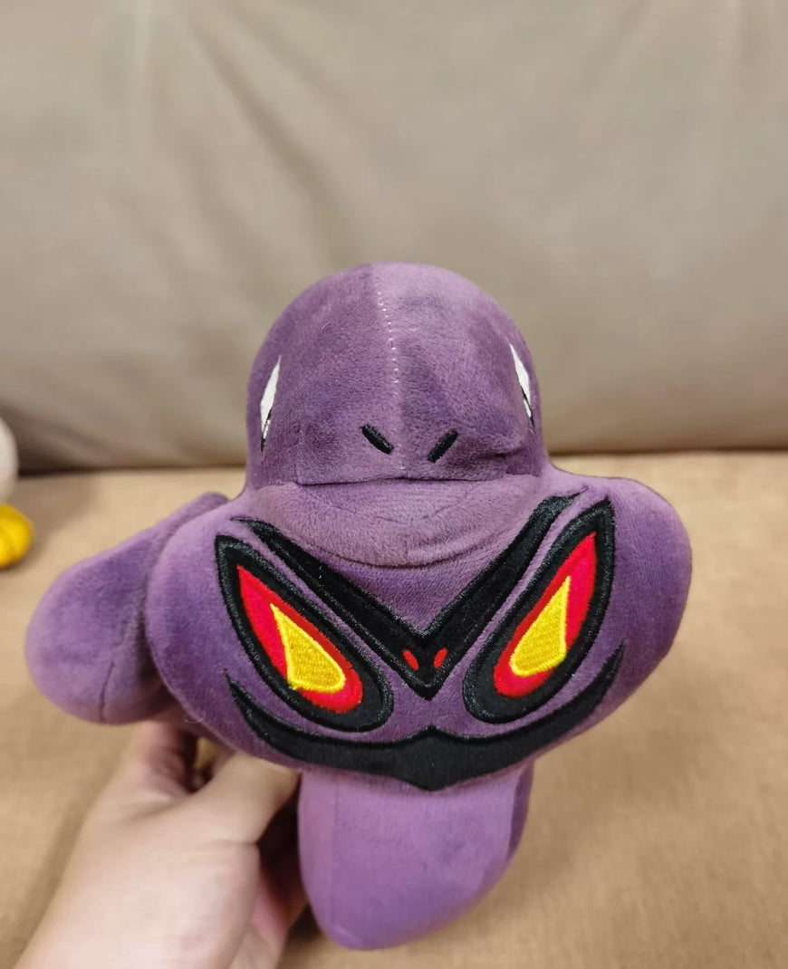 A purple plush toy resembling a cartoon monster or frog-like character, held in a person's hand against a beige couch background. The face has black embroidered details and red and yellow eye shapes.