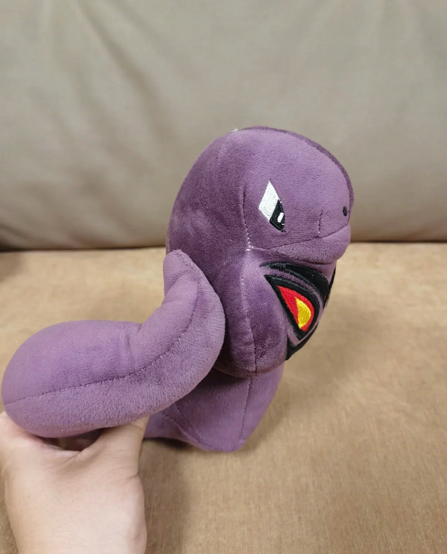 A purple plush toy resembling a snake or dragon with embroidered eyes and a red-yellow mouth detail, held by a hand against a beige fabric background.