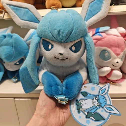 A blue and white plush toy with long ears and a small metallic keychain ring, held in a hand in front of a shelf with other plush toys. A round tag with the character illustration is attached near the bottom.