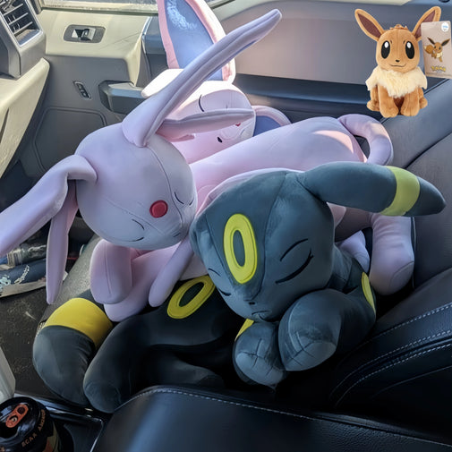A pile of plush toys featuring two large Eevee plushies and one smaller Eevee figure, arranged on a black car seat inside a vehicle.