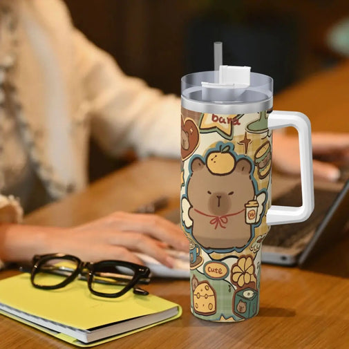 A handleless travel tumbler with a clear lid and straw, featuring a cute cartoon bear illustration, shown on a wooden desk next to a person working on a laptop.