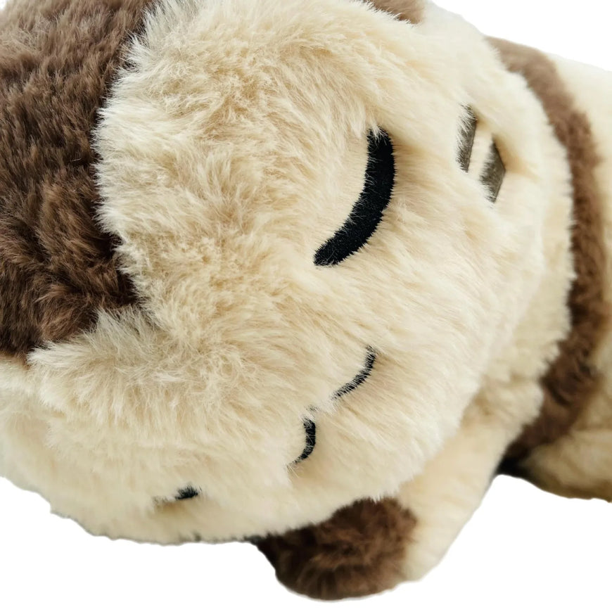 Close-up of a plush toy with soft cream-colored fur and dark brown sections, featuring embroidered black closed eyes.