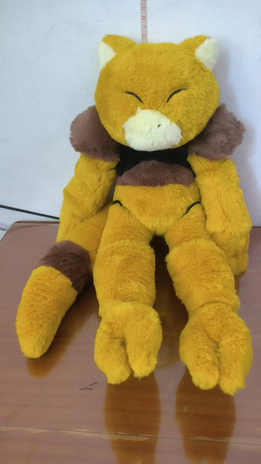A yellow plush toy with brown and cream details, resembling a stylized animal character, sitting on a wooden surface against a plain wall.