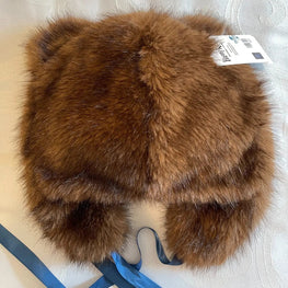 A brown faux fur trapper hat with ear flaps and blue ribbons, shown from the back on a white patterned fabric background.
