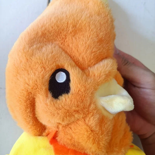 A close-up of an orange plush toy with a yellow beak and a yellow lower section, held by a person's hand against a plain light background.