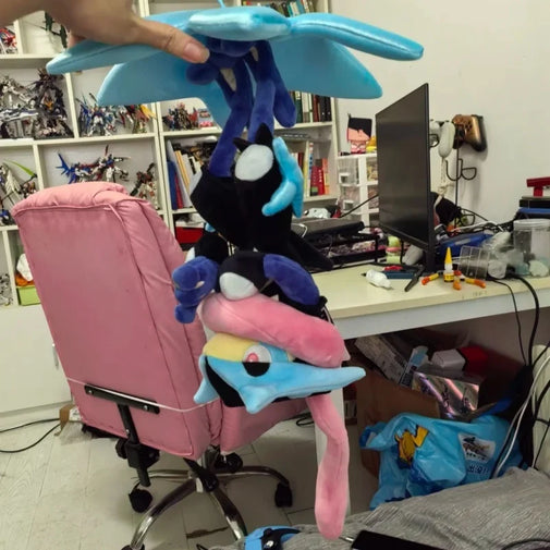A person holds a stack of plush toys, including a blue winged character, a black cat-like plush, a pink bird-like plush, and another blue/yellow plush, in a cluttered home office or playroom with a pink chair, shelves, and computer desk visible.