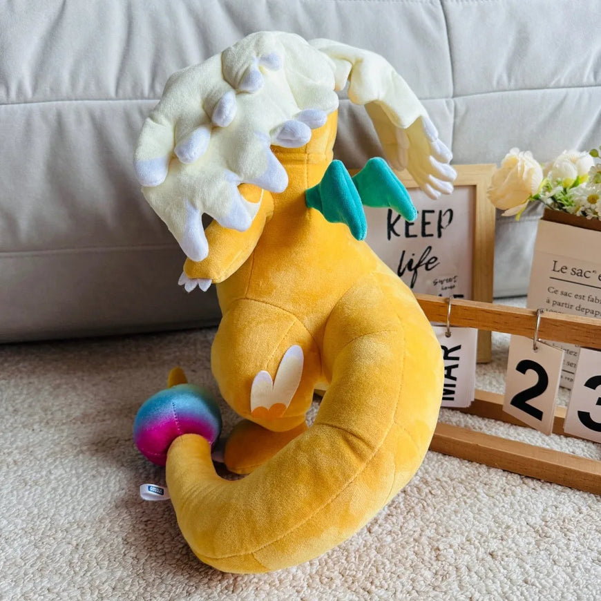 A yellow plush dragon toy with white horns and teal wings, posed on a beige carpet in front of a light gray sofa, with a small colorful ball and decorative props nearby.