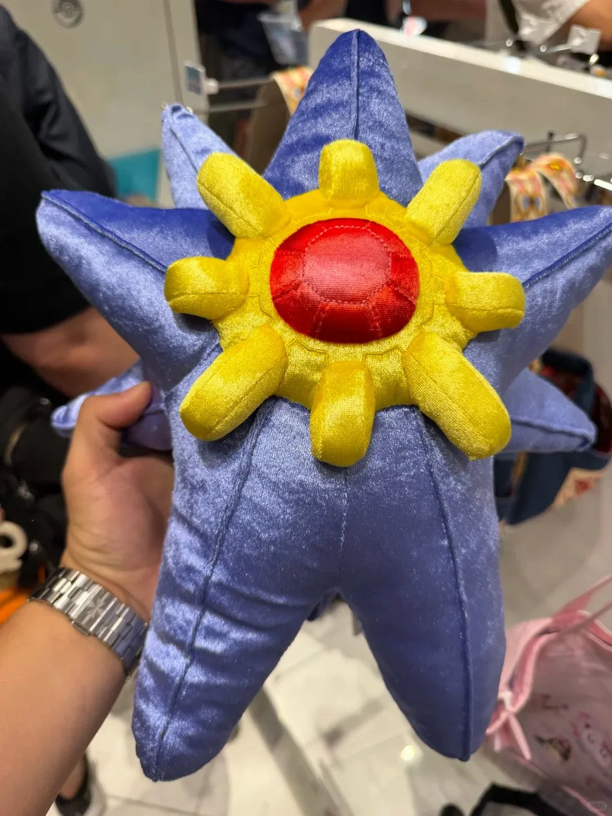 A plush starfish toy held in a hand inside a store. The starfish has a blue body, a yellow sunburst pattern, and a red center.