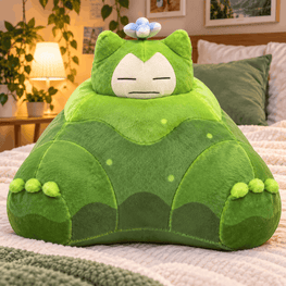 A green Snorlax plush pillow shaped like the Pokémon character, resting on a bed in a cozy bedroom setting with plants and a lamp in the background.