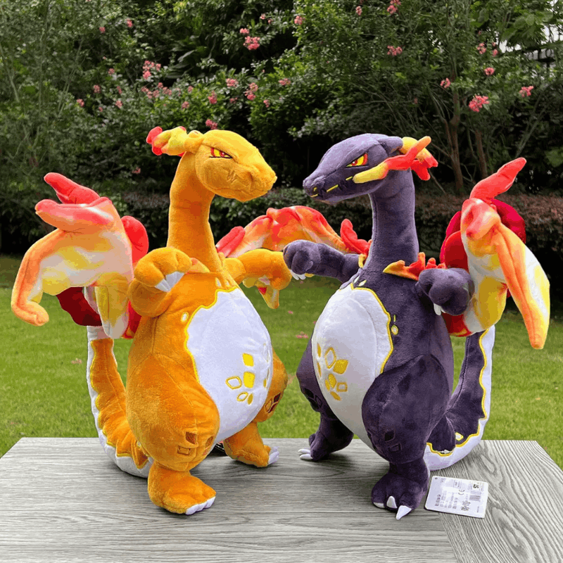 Two plush Charizard toys, one orange-yellow and one dark purple, standing on a gray wooden table outdoors with green grass and flowering bushes in the background.