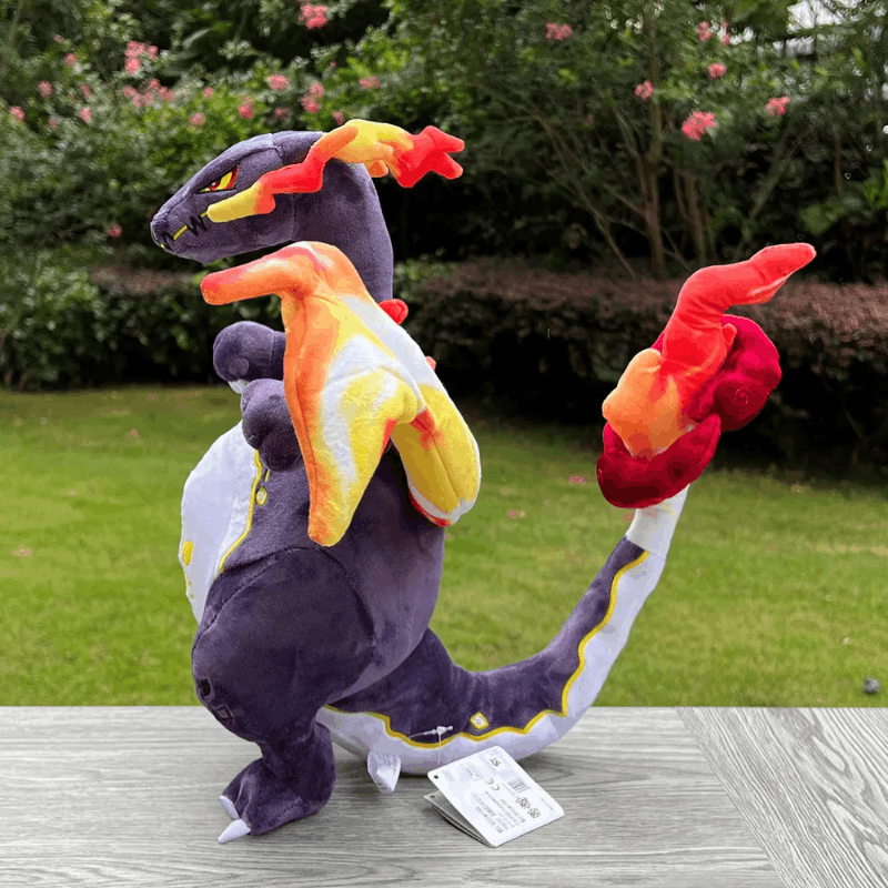 A plush toy of Charizard, the fire dragon from Pokémon, standing on a gray wooden table with a green grassy garden background. The plush features its signature dark purple body, white underbelly, and red, orange, and yellow flame patterns on its neck, wings, and tail.