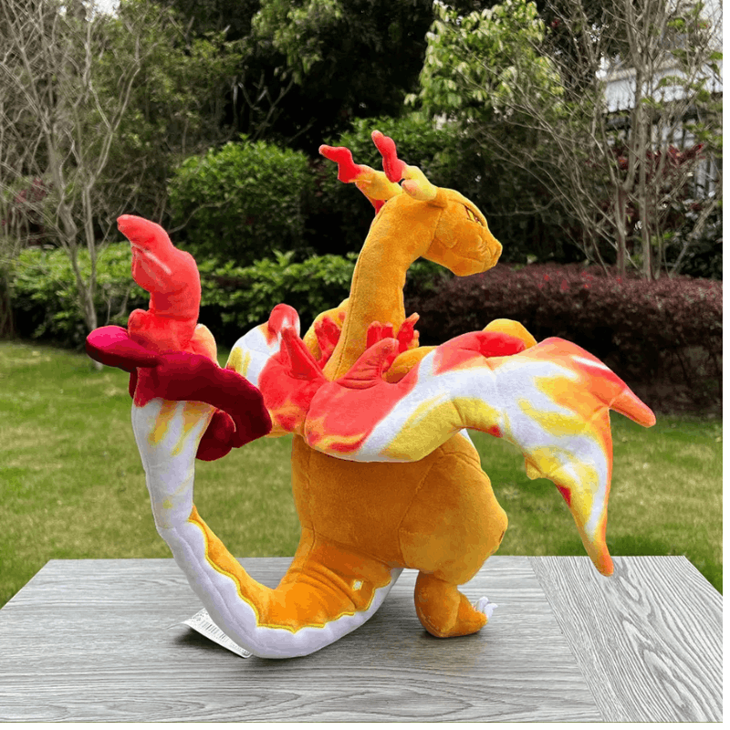 A plush toy dragon in orange, yellow, red, and white, posed on a gray wooden table with a blurred green garden background.