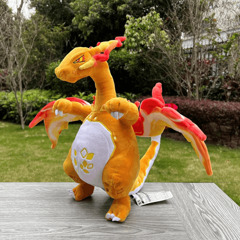 A plush toy of a fire dragon, resembling Charizard, standing on a gray wooden table with a blurred green garden background. The plush is primarily orange with a white belly, red and orange wing details, and yellow accents.