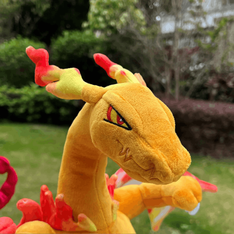 A plush toy dragon in a yellow-orange body with red and green flame-like accents, photographed outdoors on grass with blurred greenery in the background. A small watermark text is visible across the center.