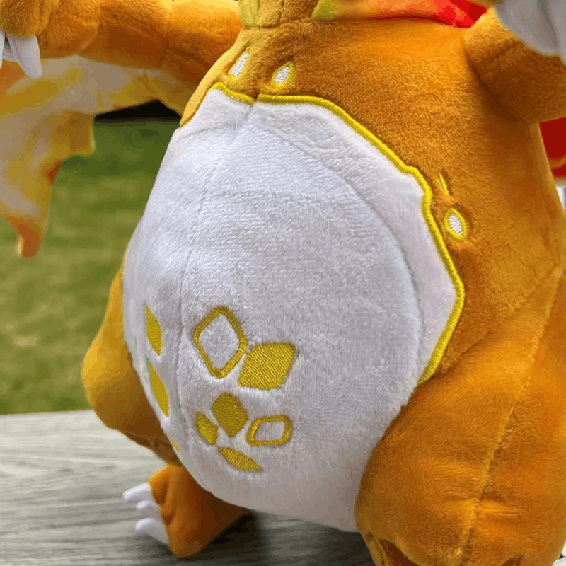 Close-up of a plush toy character with orange fur, a white belly patch, and yellow embroidered details, photographed outdoors on a wooden surface with grass in the background.
