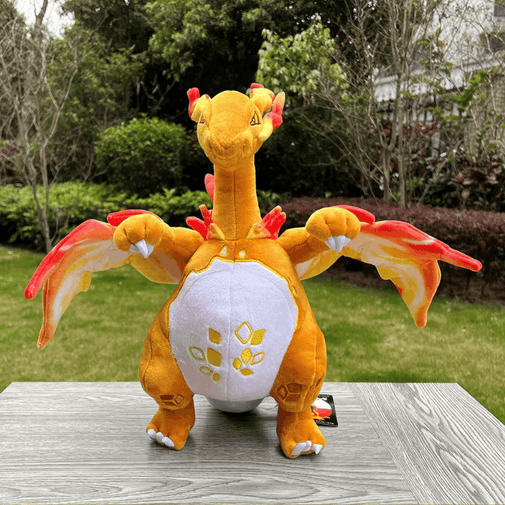 A plush toy dragon sitting on a gray wooden table outdoors, with green grass and trees in the background. The dragon is primarily orange with a white belly, red and yellow wing details, and small red horns and spikes.